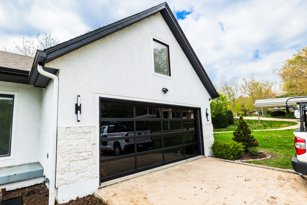 Sunny day in a neighborhood with the focus on a white house with a black garage door that needs prep for winter with garage door refurbishment in Johnson County