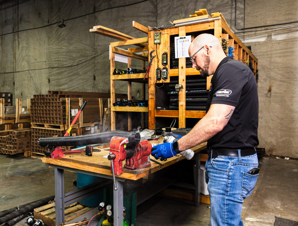 Technician working in garage at a workbench. He is wearing gloves and holding a spray can in one hand, and a garage door component in the other. This is part of a service for garage door maintenance in Johnson County.