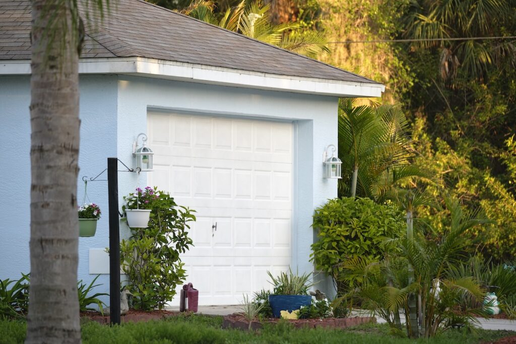 A recent white garage door installation in Shawnee on a light blue home with a garden and bushes.